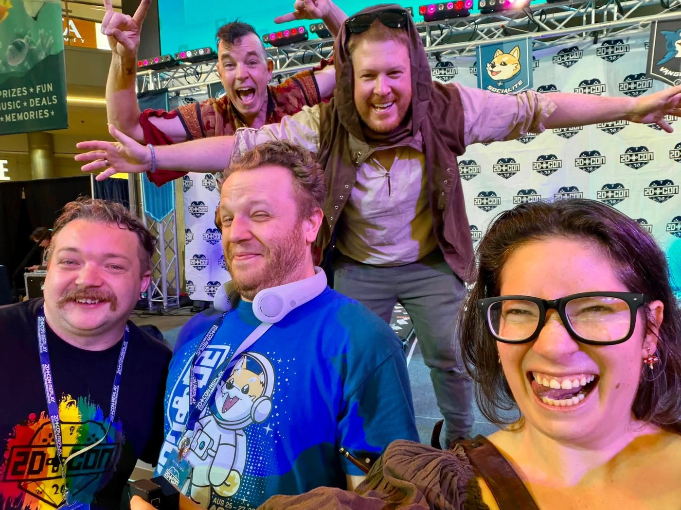 A selfie at 2D Con: band members in costume clown behind and beside two people in 2D Con shirts, in front of the convention stage.
